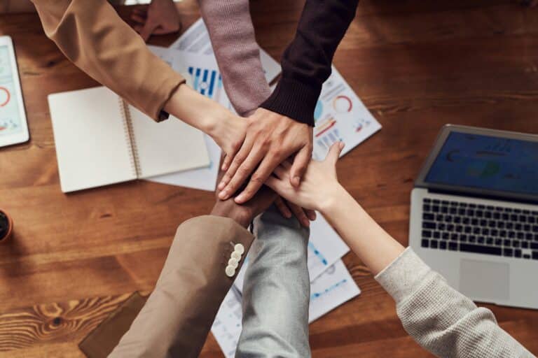 six people are leaning over an office desk and putting their hands together, united in team spirit