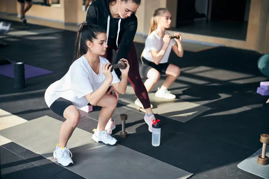Young girls are doing squats while holding dumbbells in their hands.