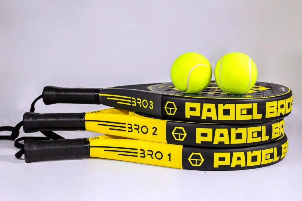 Drei Padelschläger und zwei Bälle am Boden. Padel Tennis für zwei hat sich aus Tennis entwickelt und wird schnell beliebt.